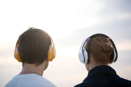 Back shot of man and woman listening to music in wireless headphones against evening cloudy sky backgroundの写真素材