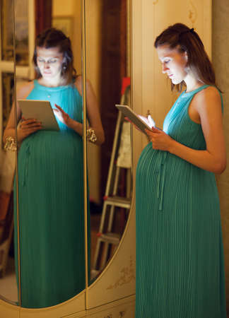 Pregnant woman in blue dress uses tablet at home standing near the sliding-door wardrobe with mirror in the eveningの写真素材