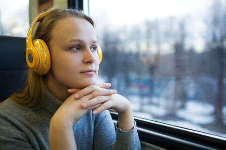 Thoughtful young woman in the train listening to music in wireless headphones and enjoying passing landscape from the windowの写真素材