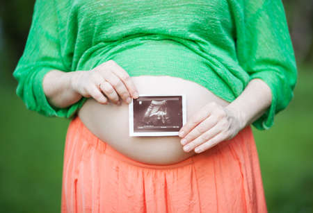 Close-up shot of a pregnant woman holding ultrasound scan in front of her bare belly. Happiness of...