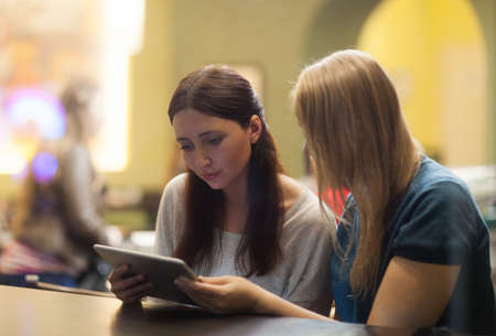 Serious blonde and brunette women sit in the restaurant watching something on the tablet