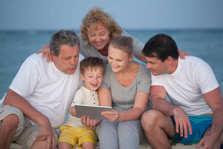 Child using tablet computer and his big family including parents and grandparents looking at screen....