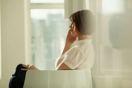 Adult woman sitting in chair and talking on phone against of window.