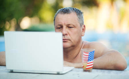 Head and Shoulders of Mature Man Soaking in Outdoor Swimming Pool and Resting on Edge of Pool with Beverage and Looking at Laptopの写真素材