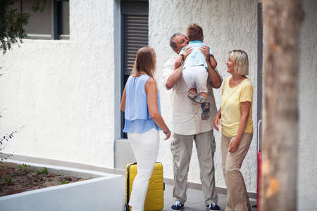 Grandfather greeting his young grandchild lifting him in the air and kissing him as he arrives on a trip with his mother outside the apartmentの写真素材