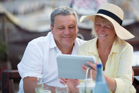 Smiling happy relaxed middle-aged couple using a tablet together on vacation as they sit at a table in a cafeteria enjoying refreshmentsの写真素材