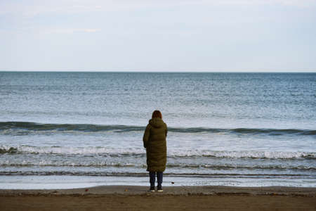 A warmly dressed woman standing at the edge of the waving sea
