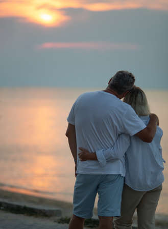 A middle-aged couple hugging each other and observing a sunset sea, we see them from the backの写真素材