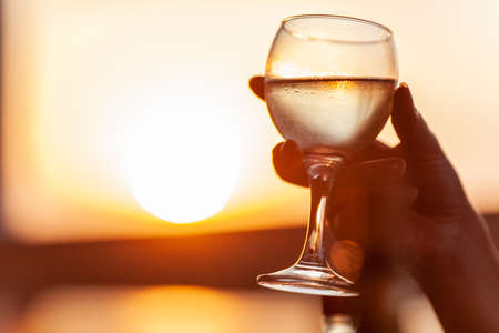 A misted glass of cold white wine in a womans hand, as gold as sunset in the backgroundの写真素材