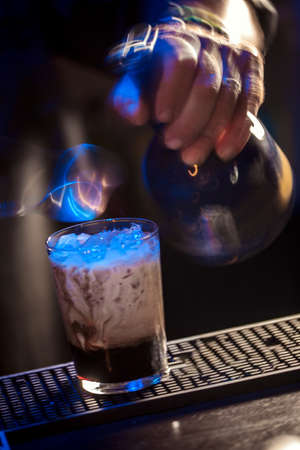 A fragment with a barmans hand, that has just finished adding a last layer into the cocktail glass