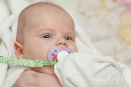 A portrait of a baby boy with grey eyes. He is holding a colorful baby dummy in his mouth