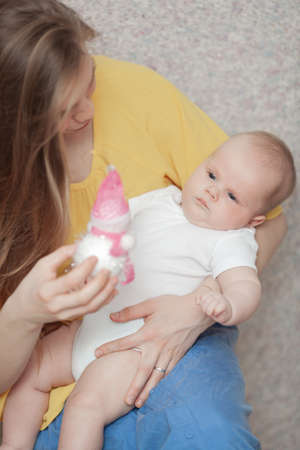 A baby boy in a white onesie is lying in arms of a long haired woman in a yellow shirt. He is...