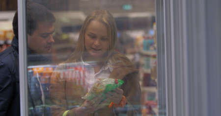 Young couple is standing by the freezer in grocery store and picking packages with frozen food.の写真素材