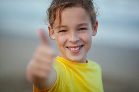 Close-up shot of a happy boy showing thumbs-up. Child with a broad smile in yellow t-shirt on blurry background. Positive thinkingの写真素材