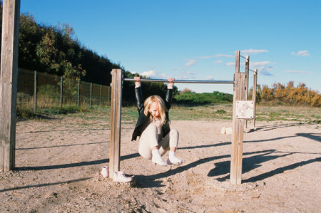 Playful little girl in leather jacket playing on sports ground while hanging on bar during weekend...