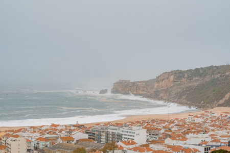 Elevated view of seaside cliffs, sandy beach, and a residential town with red-tiled roofs on a misty dayの写真素材