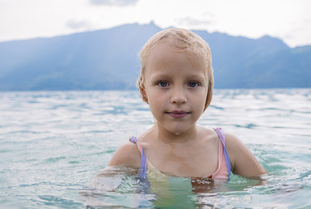 Blonde girl with blue eyes swimming in a calm lake with mountains in the backgroundの写真素材