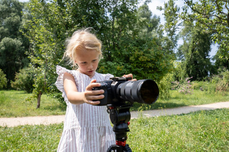 Blonde girl in a striped dress holding a professional camera mounted on a tripod in a green parkの写真素材
