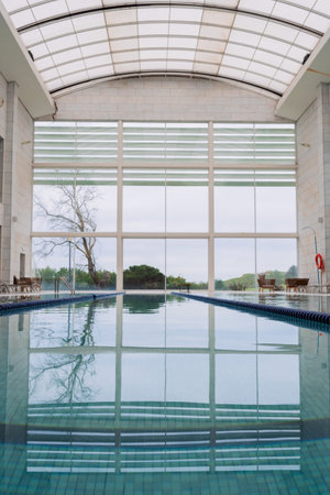 Symmetrical indoor swimming pool with lanes and a modern glass ceiling, offering a peaceful view of the surrounding greeneryの写真素材