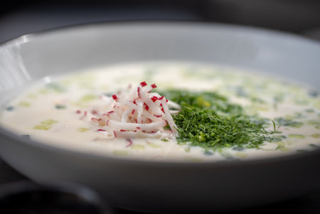 Close up of okroshka, a traditional Russian cold soup made with kefir, various chopped vegetables, and herbs, garnished with fresh dill and thinly sliced radishes, served in a white bowlの写真素材