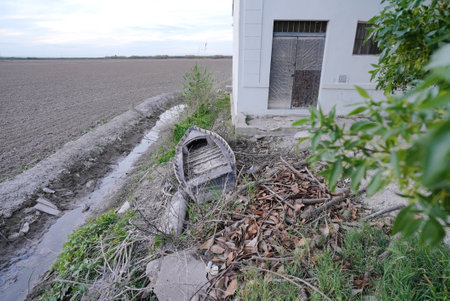 Old wooden boat abandoned on dry canal bank next to white building with rusty door and green vegetationの写真素材