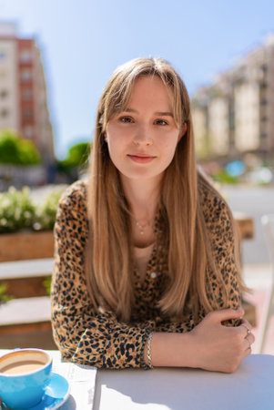 Blonde woman with long hair and a leopard print shirt sitting at an outdoor cafe table with a cup of coffee, enjoying the urban surroundingsの写真素材