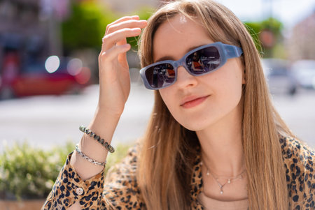 Portrait of a fashionable young woman adjusting her hair while wearing stylish sunglasses, enjoying a sunny day in an urban setting, showcasing her trendy leopard print shirt and jewelryの写真素材