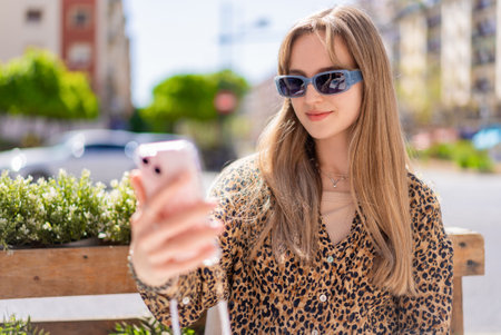 Young woman wearing sunglasses and leopard print shirt is taking selfie with her smartphone while sitting in outdoor cafe on sunny summer dayの写真素材