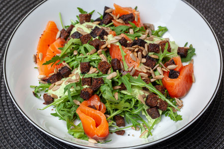 Fresh salmon salad featuring vibrant arugula, crunchy rye croutons, and sunflower seeds, all drizzled with zesty green sauce in a white bowl on a sleek black matの写真素材