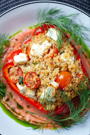 Close up of a delicious red bell pepper stuffed with couscous, feta cheese, cherry tomatoes, zucchini, and garnished with fresh dill, served on a plate with a creamy sauceの写真素材