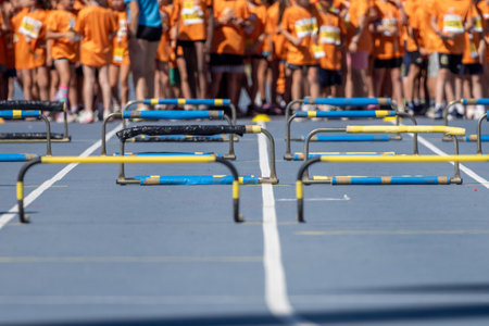 Blue athletics track with yellow and black hurdles aligned and young athletes waiting in the backgroundの写真素材