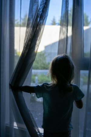 Child standing with back turned, pulling curtains aside, peering out window with wide eyed wonder,...