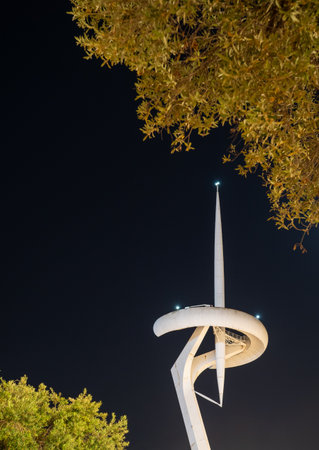 Illuminated Montjuic Communications Tower glowing against dark sky, silhouetted trees framing...