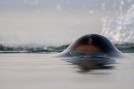 Swimmer wearing blue goggles emerging partially from water, creating rippled surface effect with...