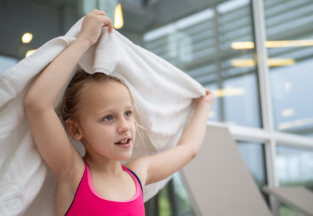 Little girl happily drying her hair with a towel after enjoying a refreshing swim in an indoor pool, radiating joy and cheerfulnessの写真素材