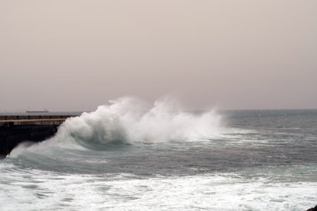 Big waves crashing against the pier during a fierce storm, with a cargo ship navigating the...