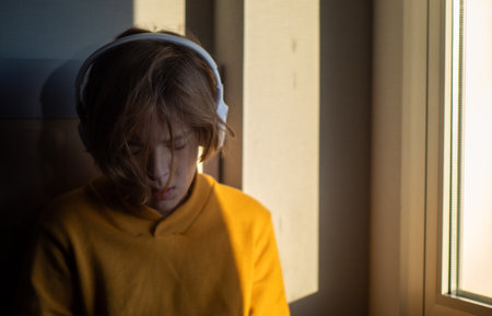 Teenager wearing headphones, immersed in music while sitting near a window, basking in the soothing...