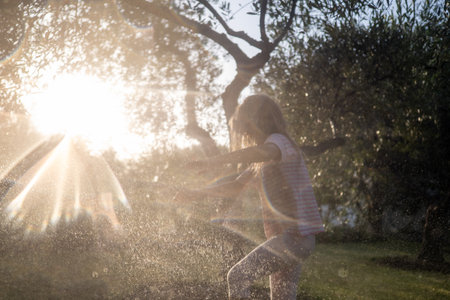 Carefree child playing joyfully with water jets in a garden during sunset, illuminated by the warm glow of golden hour lightの写真素材