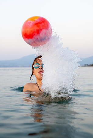 Child swimming underwater, wearing blue goggles, pushing bright inflatable ball with splashing waves in ocean backgroundの写真素材