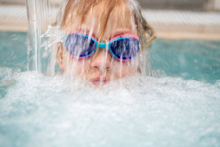 Young girl wearing swimming goggles, joyfully splashing in a sunny pool during summer vacationの写真素材