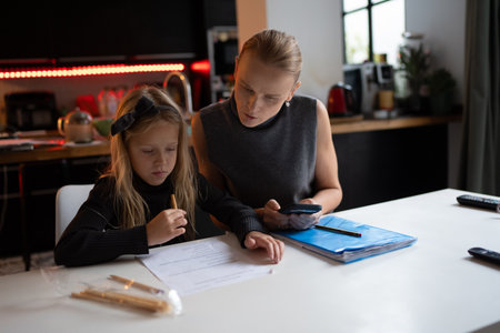 Mother assisting young daughter with homework at a kitchen table, examining worksheets and using a...