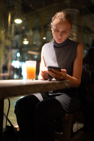 Woman sitting in a dimly lit cafe using her smartphone next to a drink on a table at night