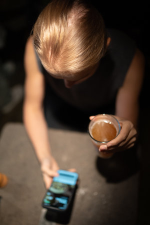 Blurred top view of a person holding a smartphone and a glass of drink in low light