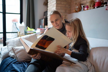 Mother and daughter sitting on a cozy sofa while looking through a large family photo album together in a warm living room settingの写真素材