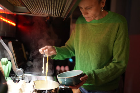 Woman lifting cooked pasta from pot with fork while standing in modern home kitchen