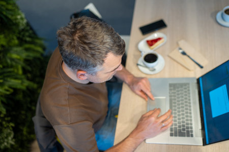 Overhead view of a man using a laptop at a table with coffee and a slice of cake filmed from the...
