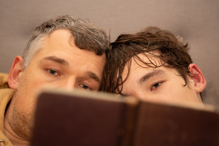 Father and son lying close reading a book with only their eyes visible focused on the pagesの写真素材