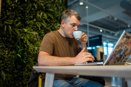 Man sits at a table with a laptop and drinks coffee in a modern indoor space with plantsの写真素材