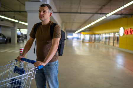 Man with backpack pushing an empty shopping cart in a dimly lit parking garage with a yellow wall...