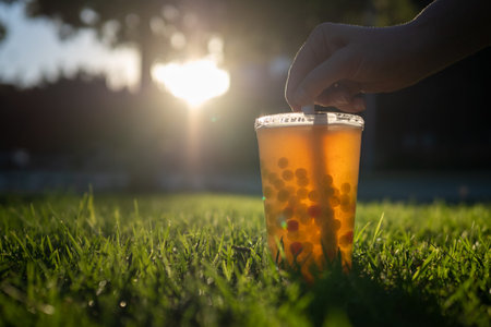 Person hand reaching for straw in cup of bubble tea standing on green grass at sunset